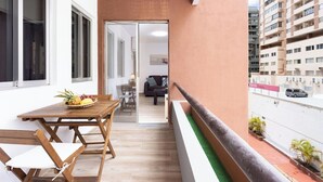 Uma varanda com mesa e cadeiras de madeira, vista para a rua e um edifício ao fundo.