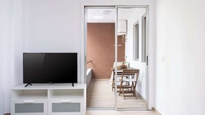 Uma sala de estar moderna com televisão de tela plana em um móvel branco, um espaço de jantar com mesa e cadeiras, e uma porta de vidro deslizante que dá para uma varanda.