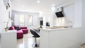 A modern kitchen with white cabinetry, a black countertop, and a built-in stove. There is a pink sofa and a black bar stool in the adjacent living area.