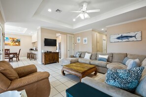 A spacious living room with a brown leather sofa, a wooden coffee table, and a beige couch with patterned cushions. A ceiling fan and a flat-screen TV are mounted on the wall.