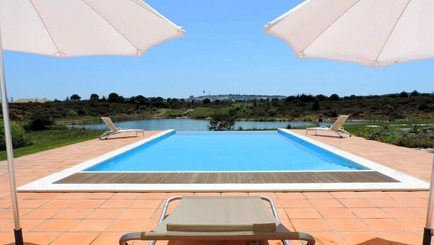 Ein rechteckiger Swimmingpool mit zwei weißen Sonnenschirmen und Liegestühlen auf einer gefliesten Terrasse, mit Blick auf einen See und die Ferne.