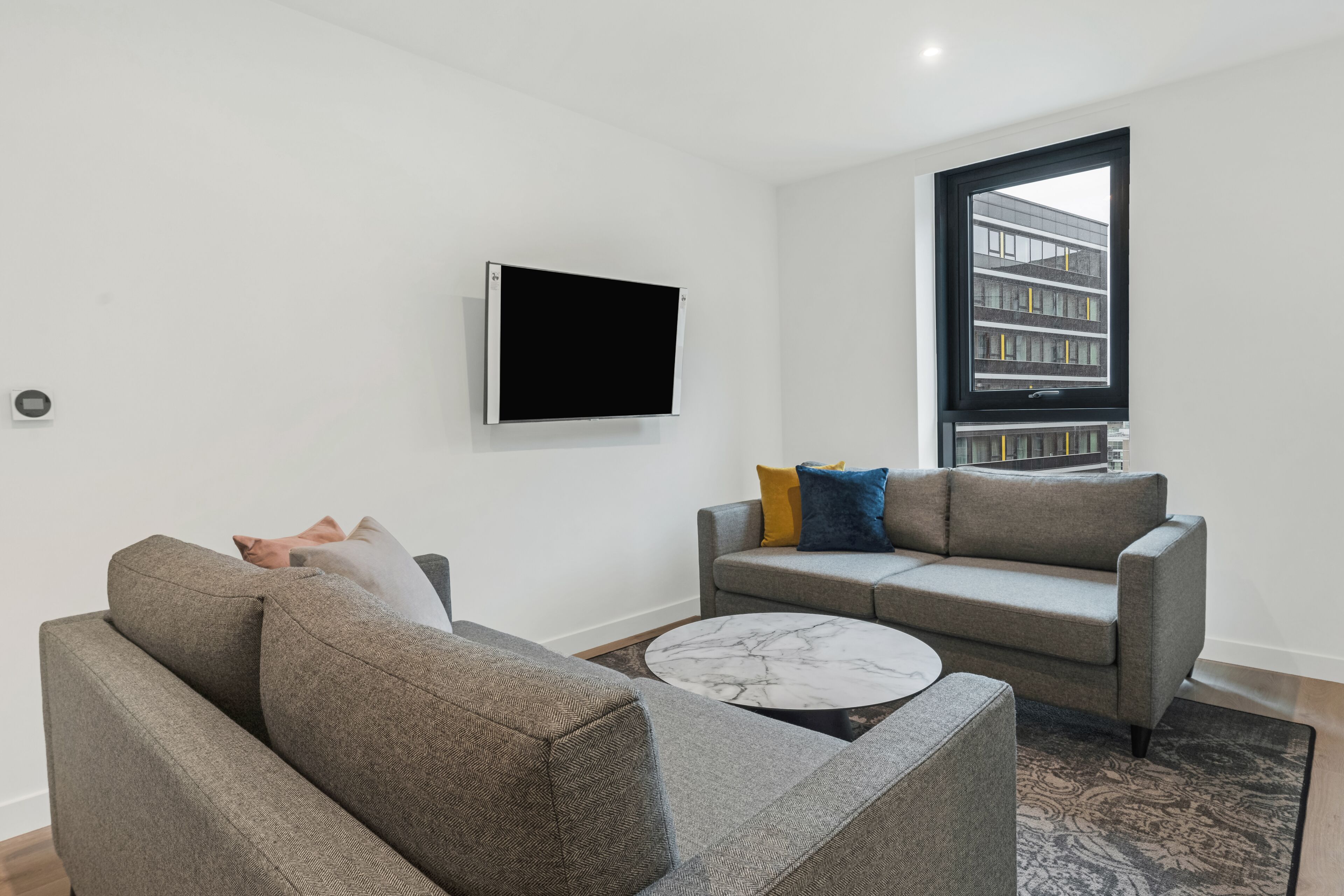 A modern living room with two grey sofas, a flat-screen TV mounted on the wall, and a large window with a view of a building.