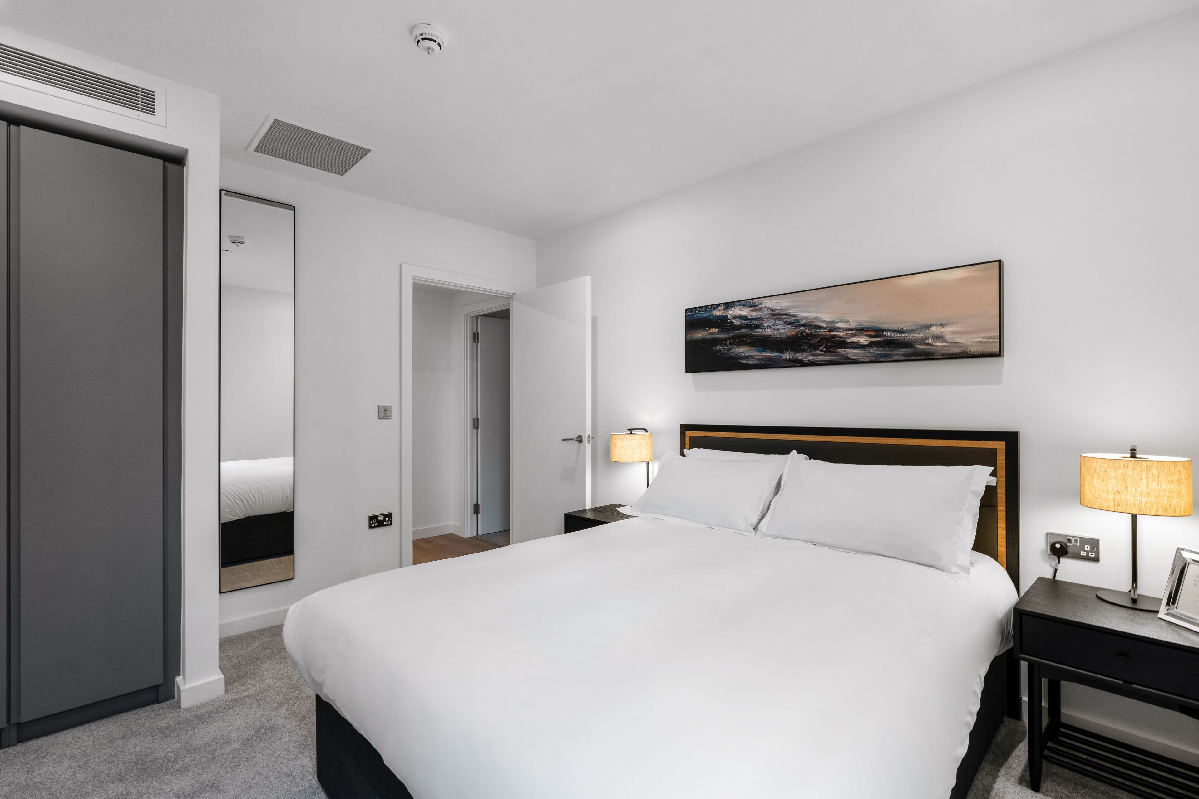 A modern hotel room with a large bed, a nightstand with a lamp, a wall-mounted painting, and a closet.