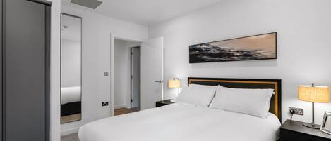 A modern hotel room with a large bed, a nightstand with a lamp, a wall-mounted painting, and a closet.