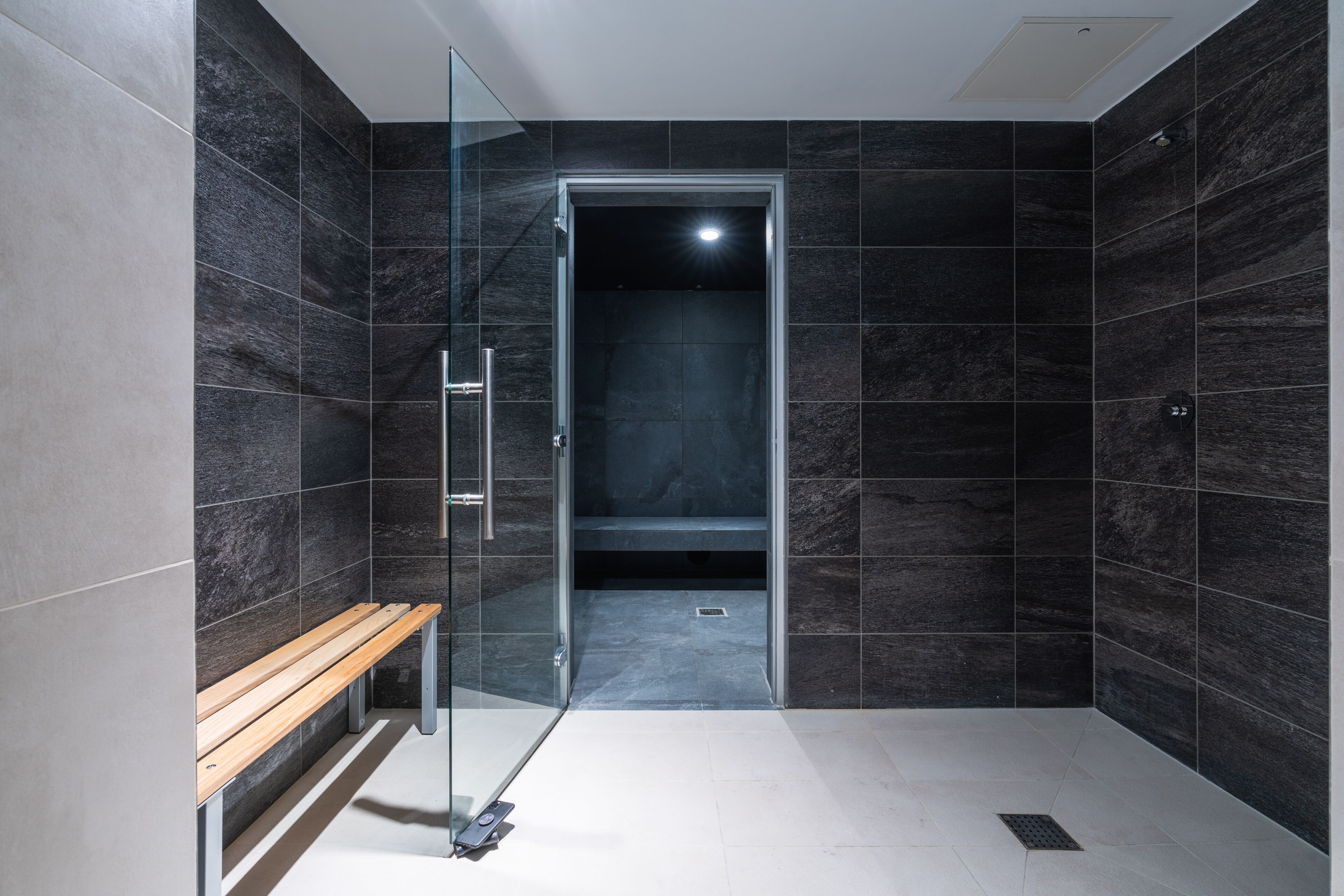 A modern bathroom with dark tiles, a glass shower enclosure, and a wooden bench.