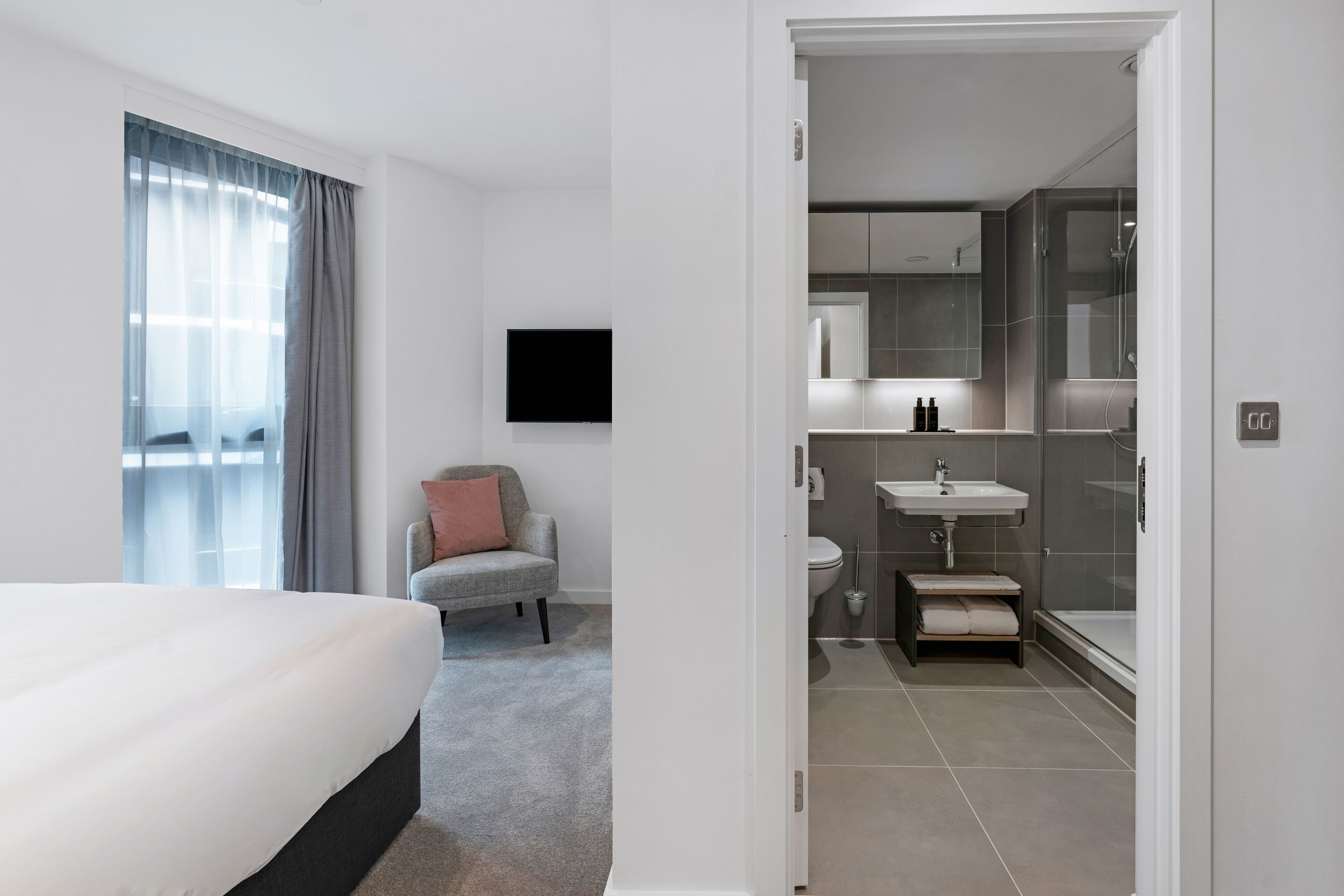 A modern hotel room with a large bed, a grey armchair, a flat-screen TV, and a bathroom with a glass shower enclosure.