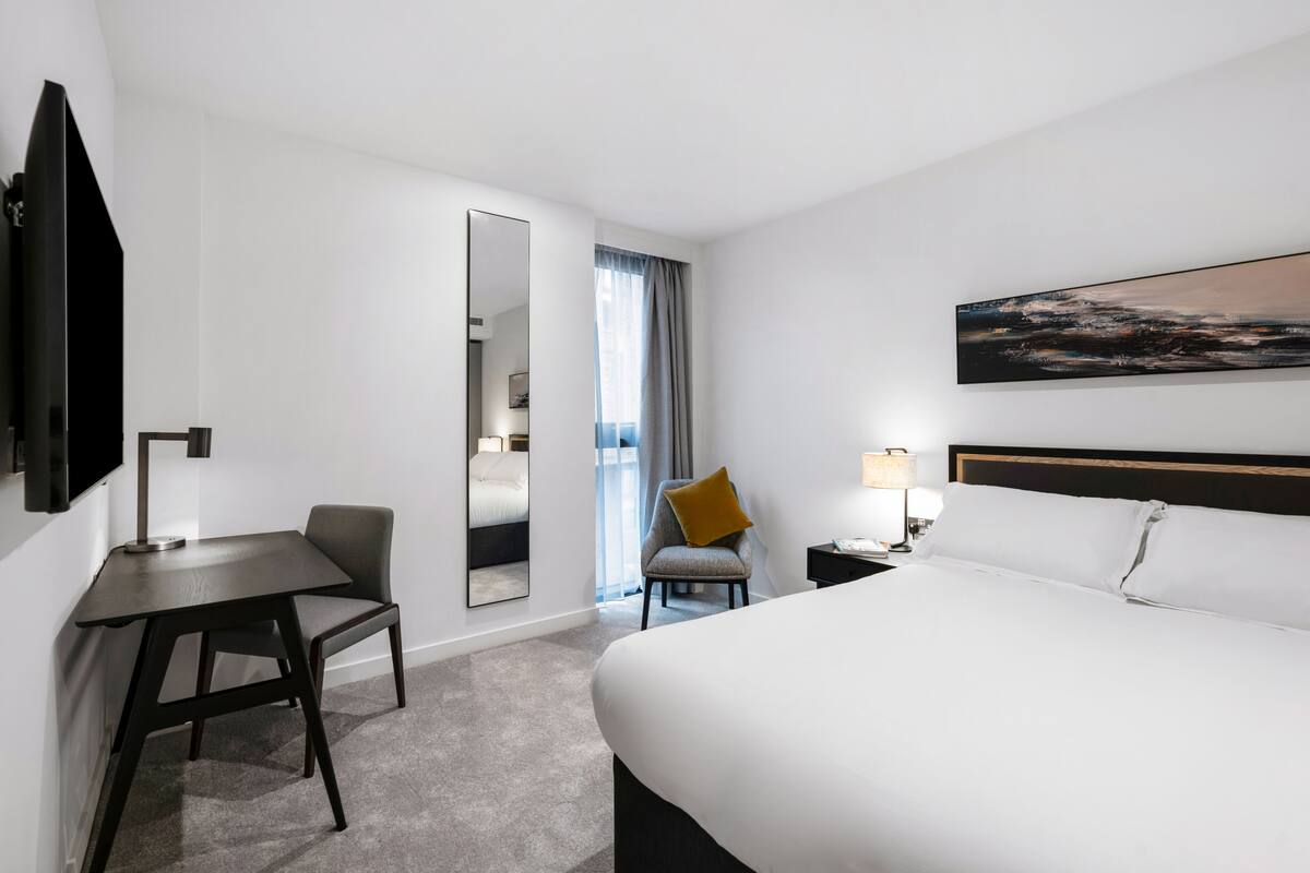 A hotel room with a large bed, a desk, a chair, a TV, and a painting on the wall.