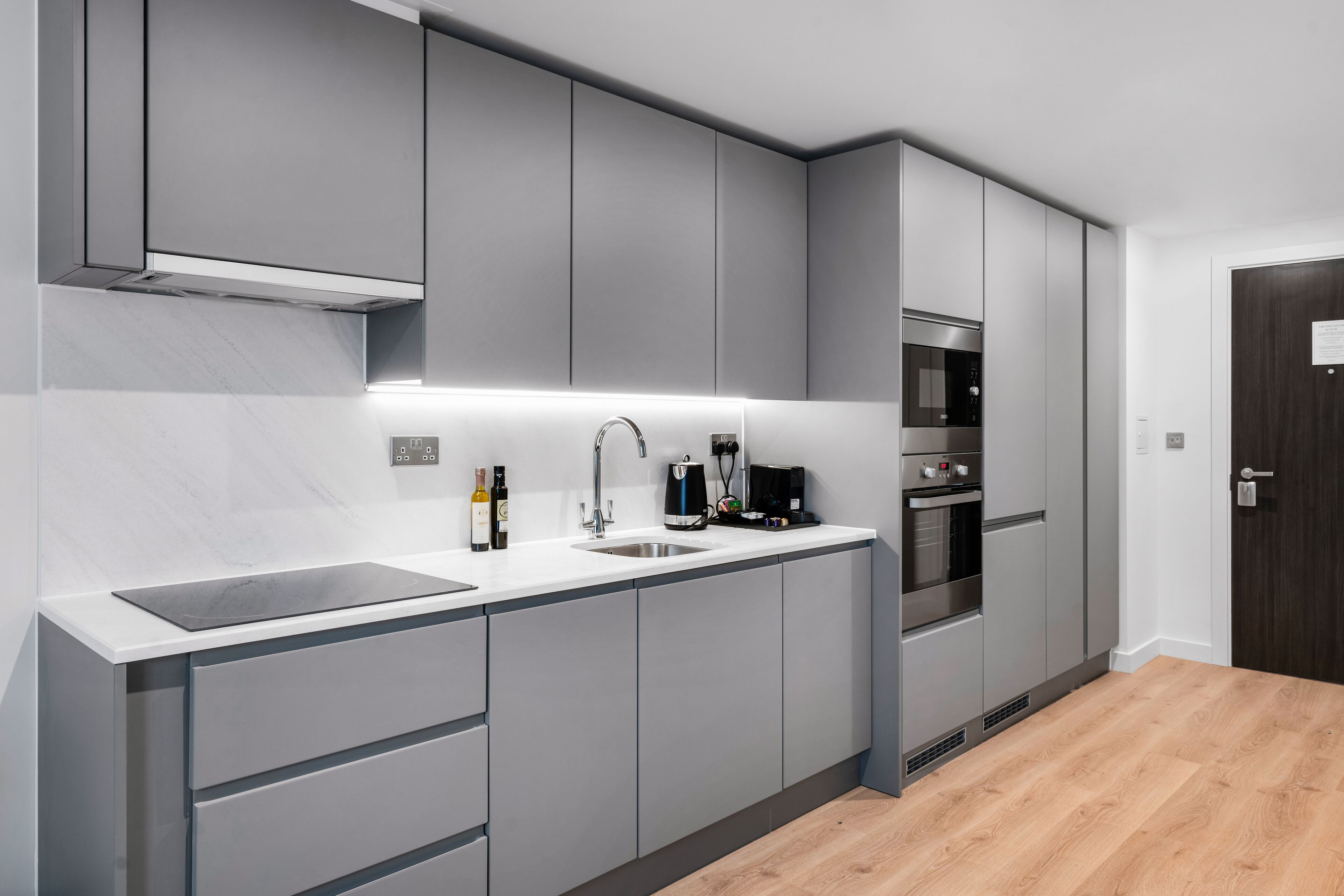 A modern kitchen with grey cabinets, a white countertop, a built-in oven, and a stainless steel sink.