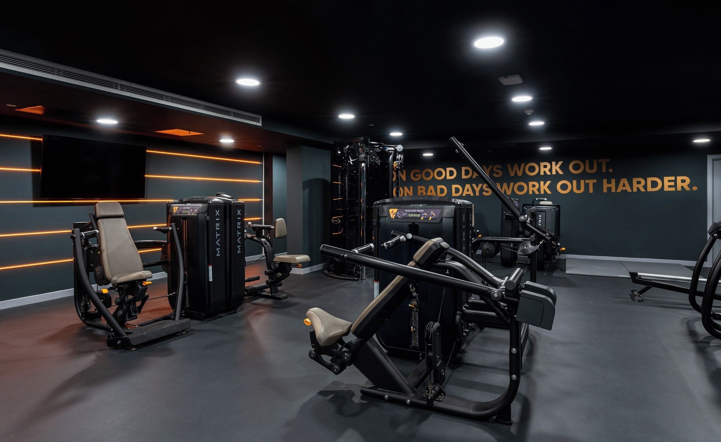 A modern gym with various exercise machines, a motivational quote on the wall, and a dark color scheme.