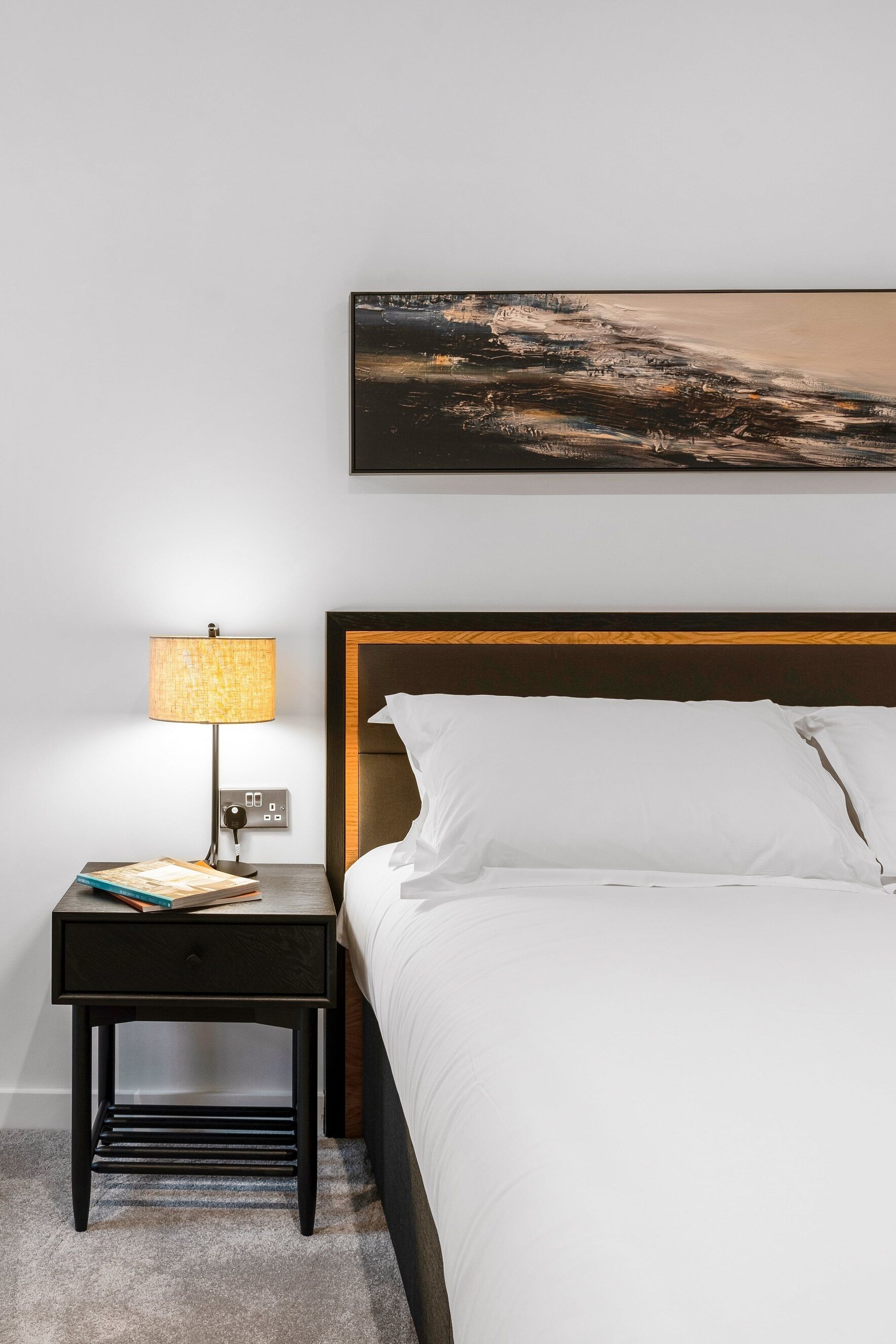 A neatly made bed with white linens, a wooden headboard, and a bedside table with a lamp and a book.