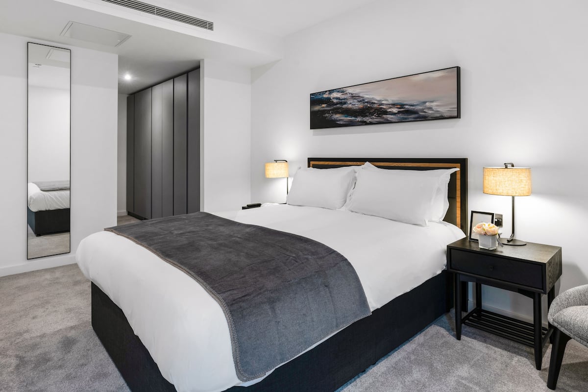 A modern hotel room with a large bed, bedside lamps, a painting on the wall, and a mirror.