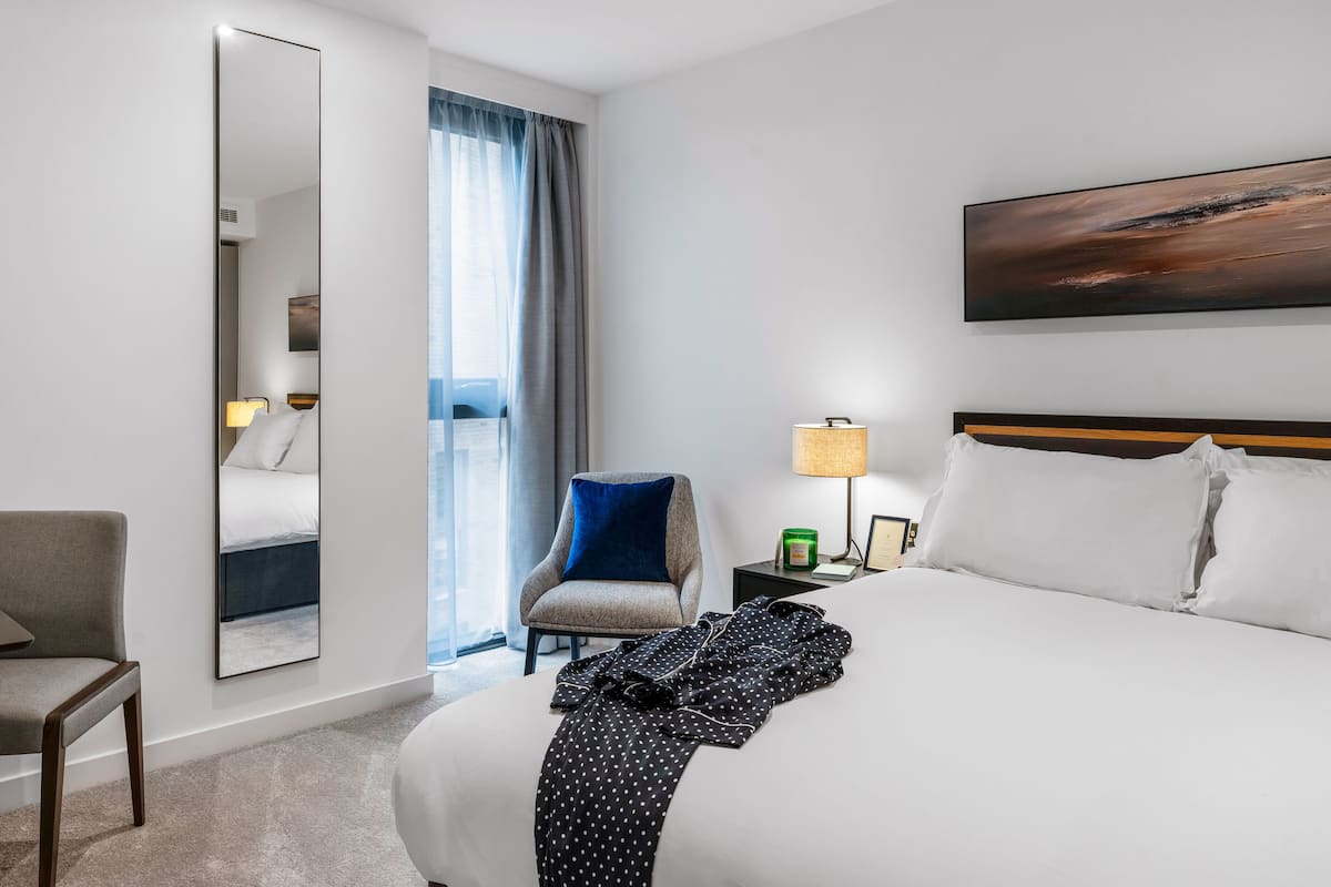 A hotel room with a large bed, a chair, a desk, a mirror, a painting, and a view of the outdoors.