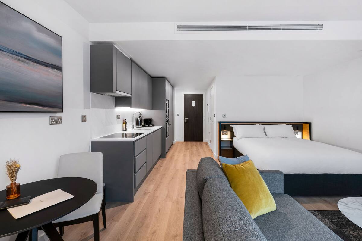A compact hotel room with a kitchenette, a sofa with a yellow pillow, a bed with white bedding, and a painting on the wall.