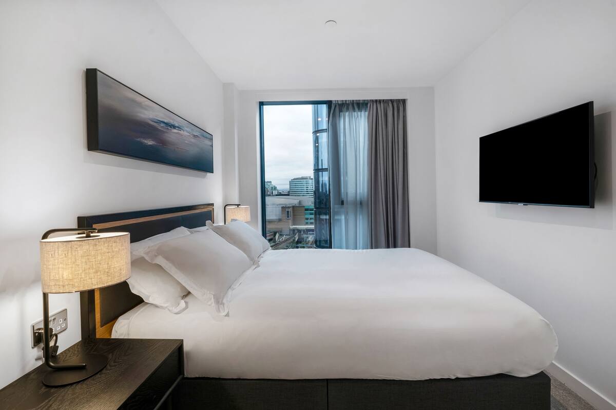 A modern hotel room with a large bed, a flat-screen TV, a nightstand with a lamp, a painting on the wall, and a view of the city through a large window.