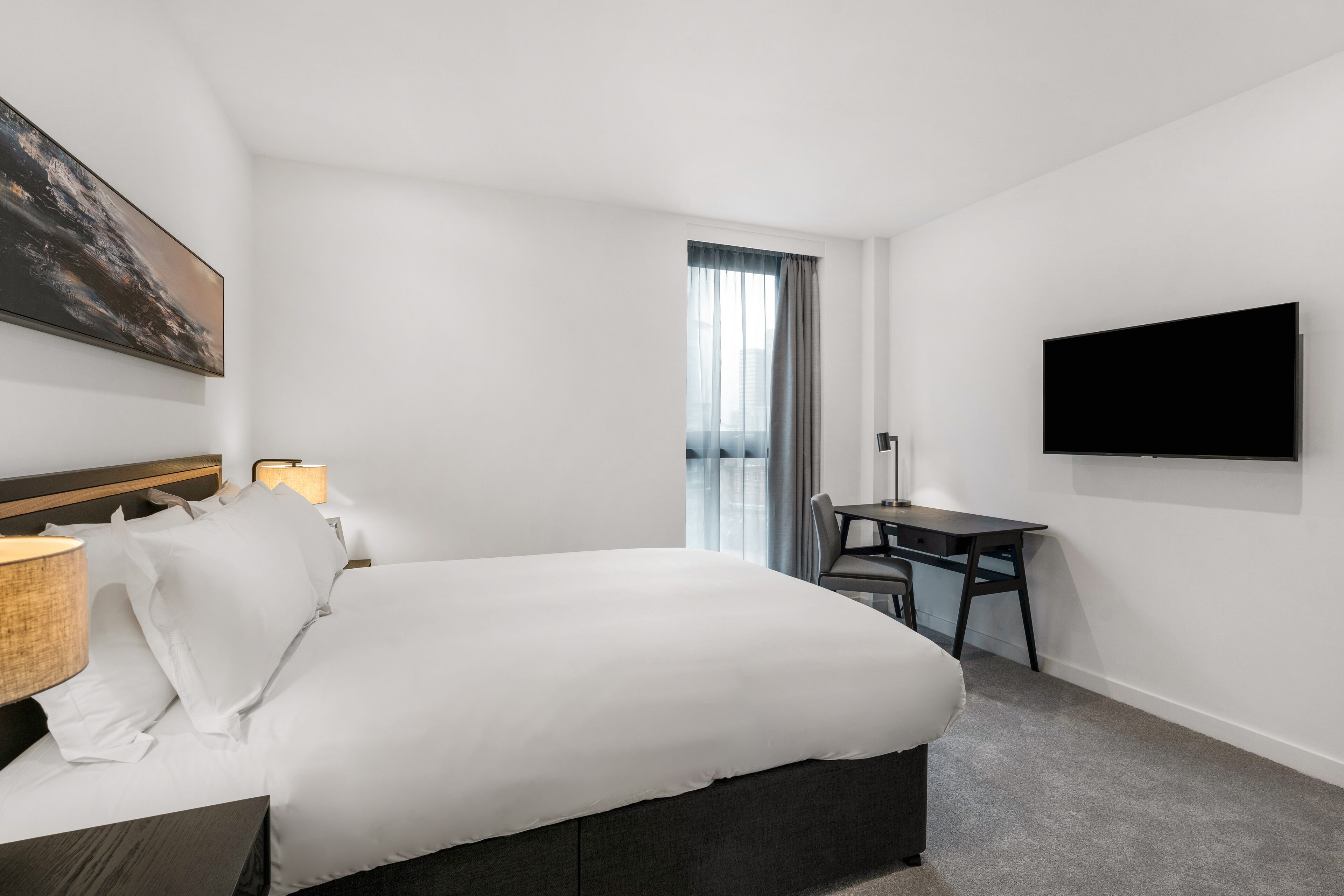 A hotel room with a bed, desk, chair, TV, and artwork on the wall.