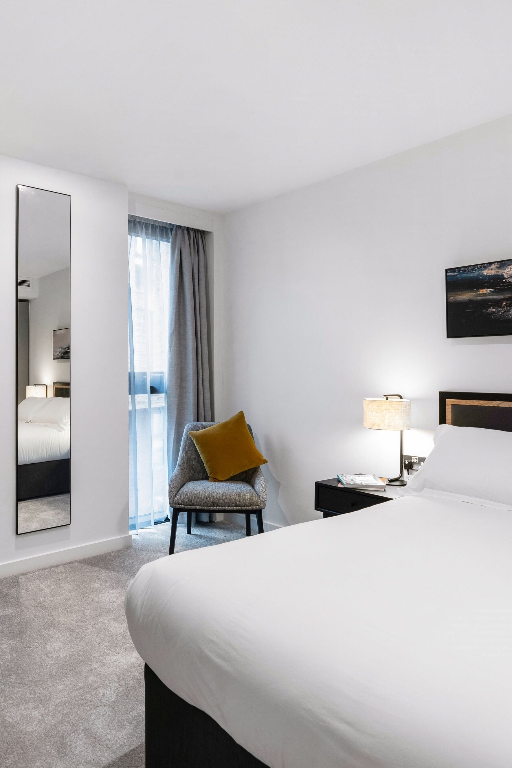 Een hotelkamer met een groot bed, een stoel met een geel kussen, een nachtkastje met een lamp, een spiegel en een raam met gordijnen.