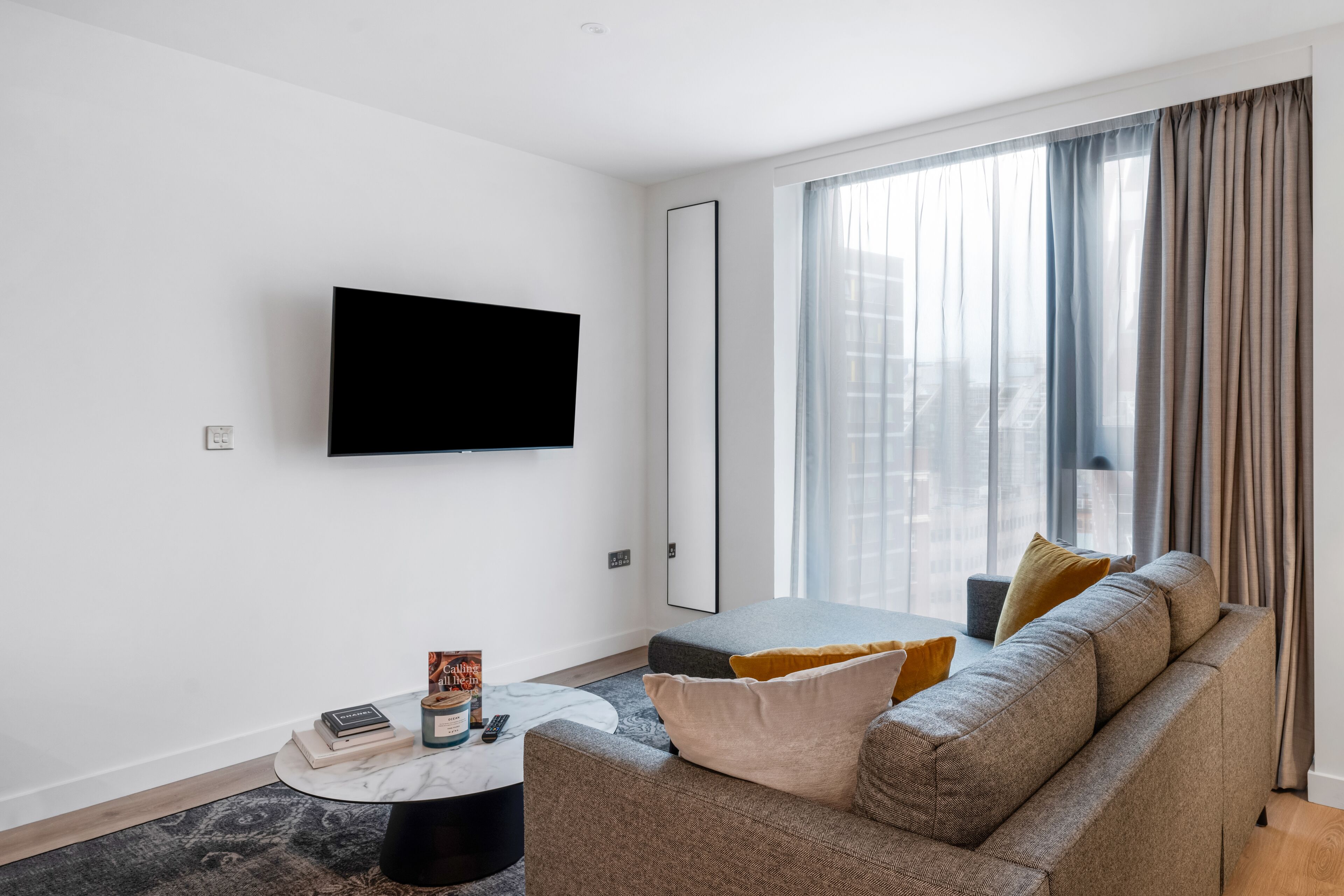 A modern living room with a large flat-screen TV mounted on the wall, a grey sofa with cushions, a round coffee table, and a large window with curtains.