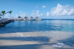 Une plage à l’eau d’un bleu limpide, avec un rivage sablonneux et une rangée de constructions aux toits de chaume.