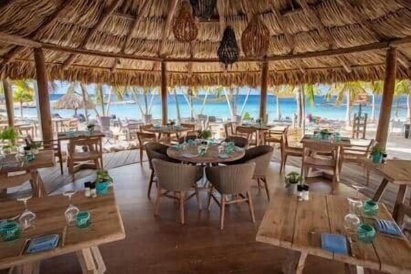 An outdoor dining area with tables and chairs, a thatched roof, and a view of the ocean.