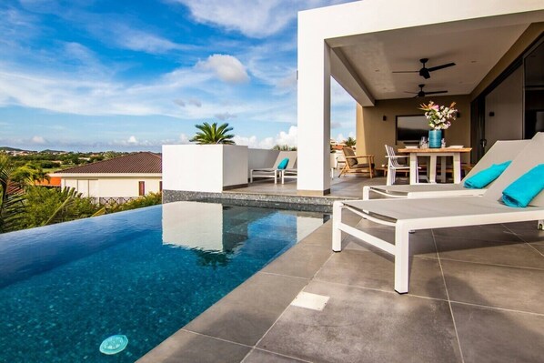 Un área de piscina moderna al aire libre con sillones de descanso y una mesa, rodeada por un edificio blanco con vistas al paisaje.
