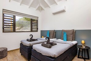 Ein Zimmer mit zwei Betten, einem Fenster mit Jalousien, einer Klimaanlage und einem kleinen Tisch mit einer Lampe.