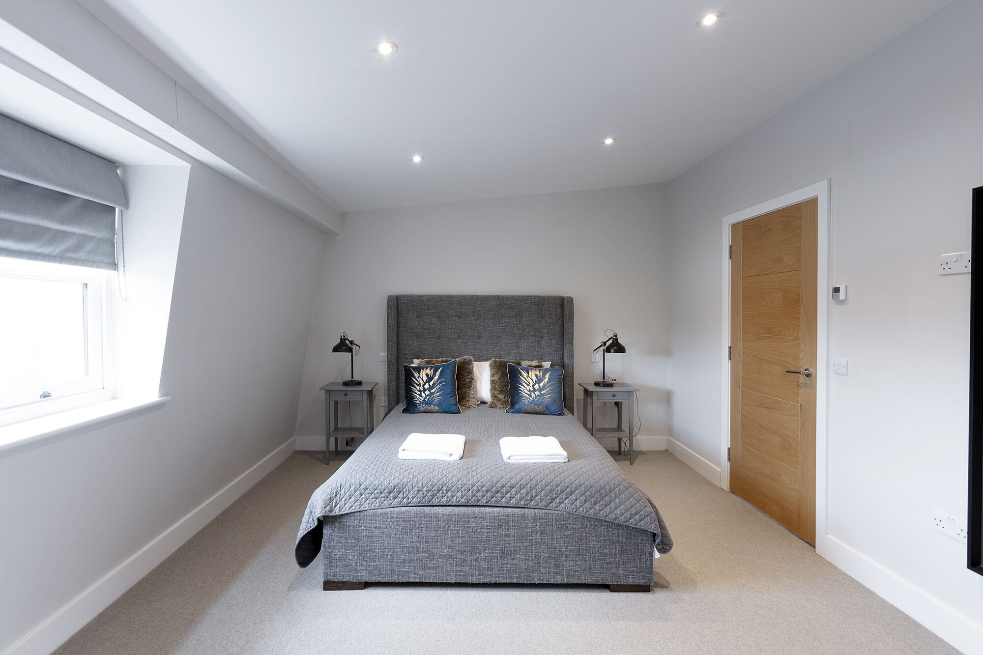 A bedroom with a large bed, two bedside tables, a window with blinds, a wooden door, and a ceiling light.