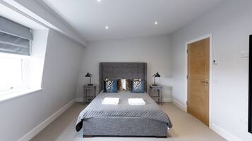 A bedroom with a large bed, two bedside tables, a window with blinds, a wooden door, and a ceiling light.