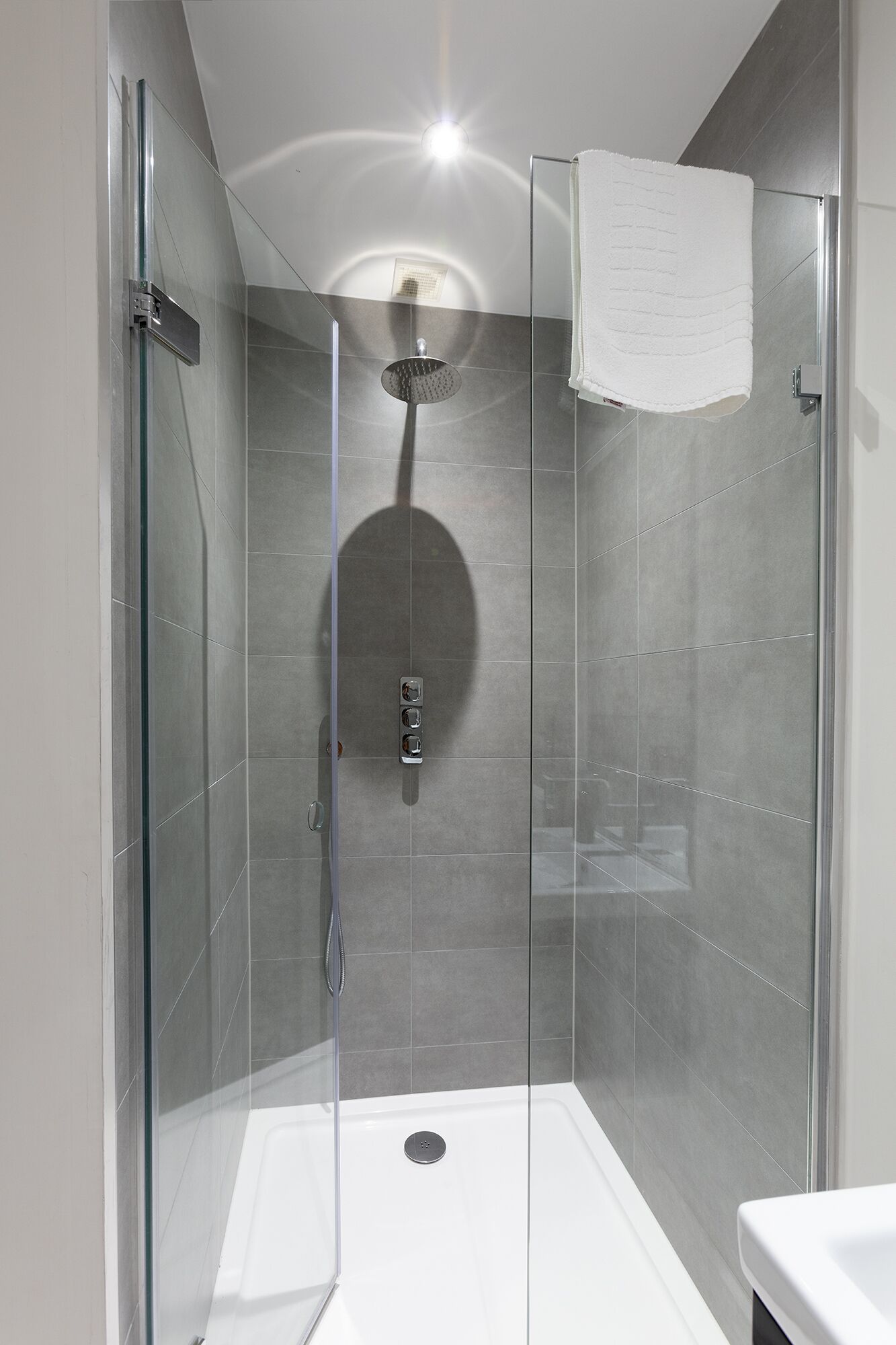 Una ducha moderna con puerta de cristal, un cabezal de ducha de mano y una toalla blanca colgada en la pared.