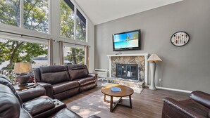 A living room with a leather sofa, a stone fireplace, a television, and a view of trees through large windows.