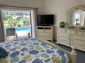 Une chambre à coucher comprenant un lit, une télévision, une commode et une grande fenêtre donnant sur une piscine et un espace verdoyant.