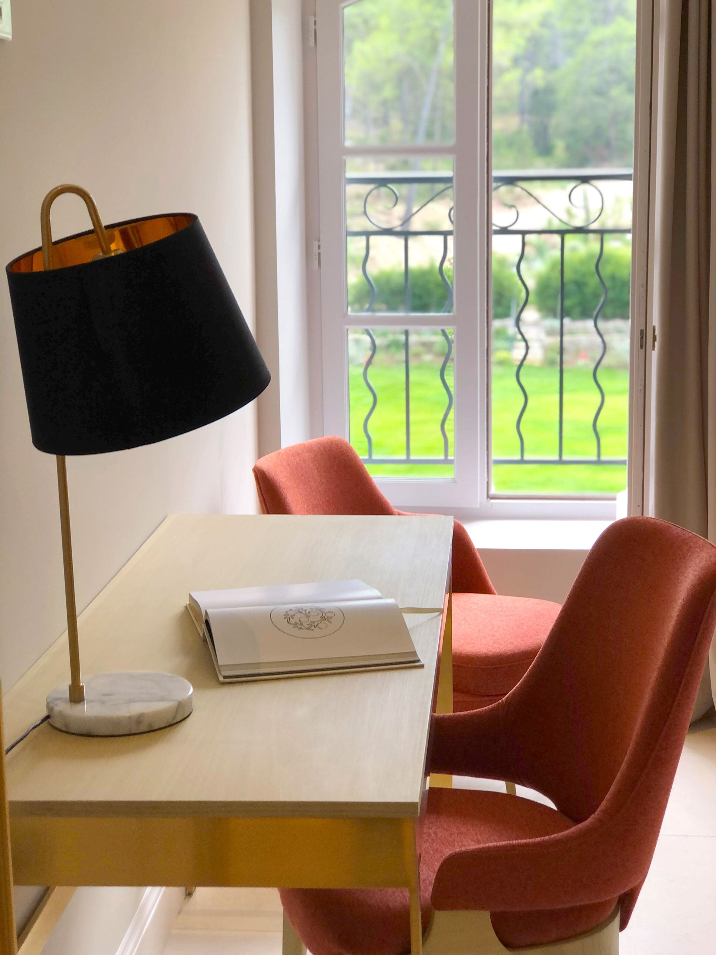 A room with a table, a lamp, a chair, and a view of a balcony with greenery.