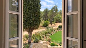 An open window with a view of a landscaped garden, including a tree, a lawn, and a stone pathway. - Domaine de Valmouriane (St.-Rémy-de-Provence)