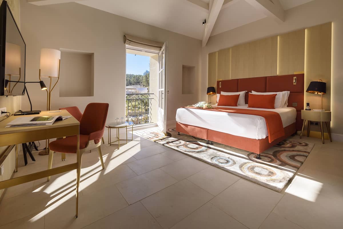 A modern hotel room with a large bed, a desk with a chair, a balcony, and a view of the outdoors.
