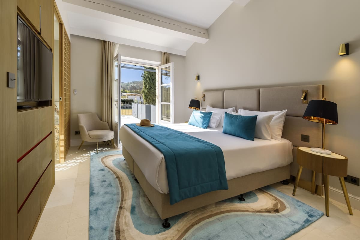 A bedroom with a bed, bedside lamps, a chair, a TV, and a view through an open door.