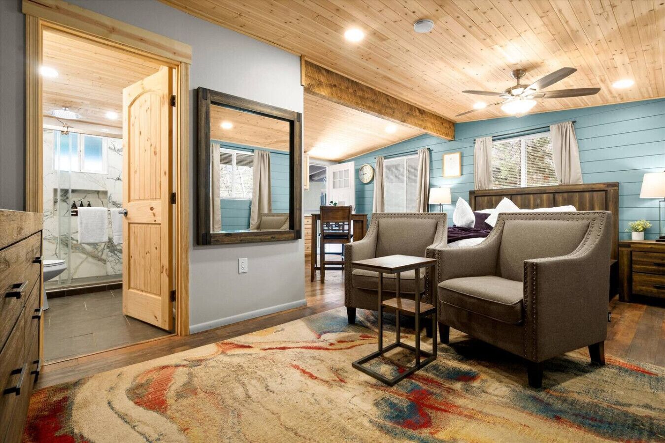 A spacious room with a large mirror, wooden ceiling, and a ceiling fan. There is a cozy seating area with two armchairs and a small table.