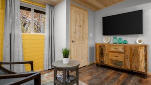 Une pièce avec un plafond en bois, un grand téléviseur fixé au mur, un meuble en bois orné d’objets décoratifs et une fenêtre avec des rideaux.