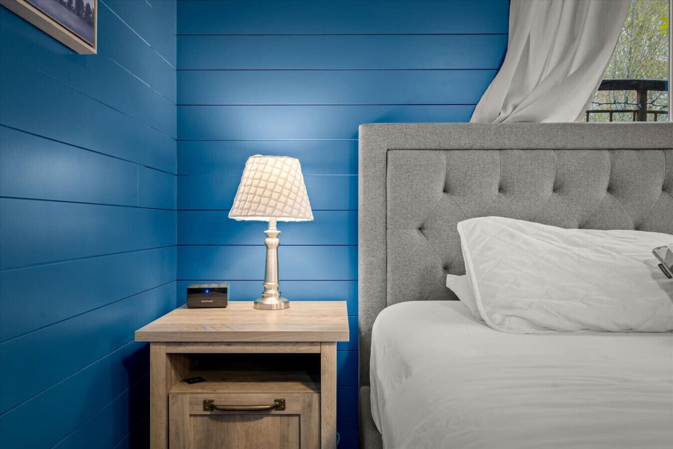 Un dormitorio con pared azul, una mesita de noche con lámpara, una cama con cabecero gris y una ventana con cortina.