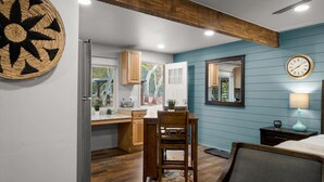A compact kitchen with wooden cabinets, a dining area with a chair, a clock on the wall, and a window with a view of trees. - 15 Creekside Retreat (Sedona)