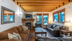 Un salon chaleureux avec une cheminée en pierre, des canapés en cuir, des poutres en bois et de grandes fenêtres donnant sur l’extérieur.