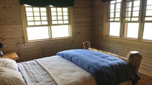 A wooden cabin room with a bed, a nightstand, and two windows with curtains.