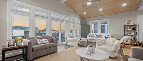 A spacious living room with a high ceiling, large windows, and a view of the sunset.