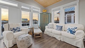 A spacious living room with two white sofas, a wooden coffee table, and a patterned ottoman. Large windows offer a view of the outside. - Gemini Dream #102 - Soundfront w/ Pool, Boat Dock, Elevator, Dogs Allowed! (Topsail Beach)
