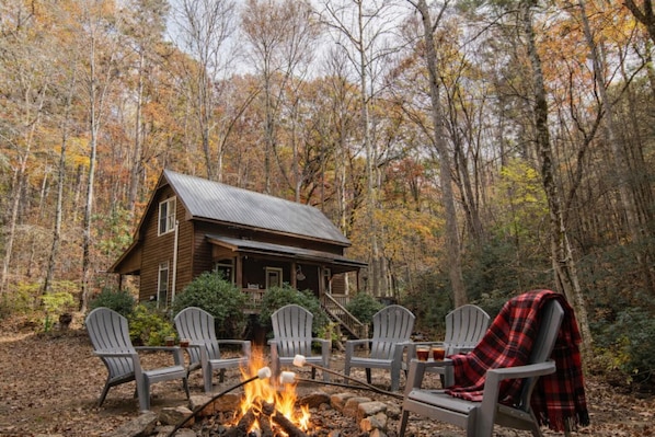 A cabin with a fire pit, surrounded by Adirondack chairs, set in a forest.
