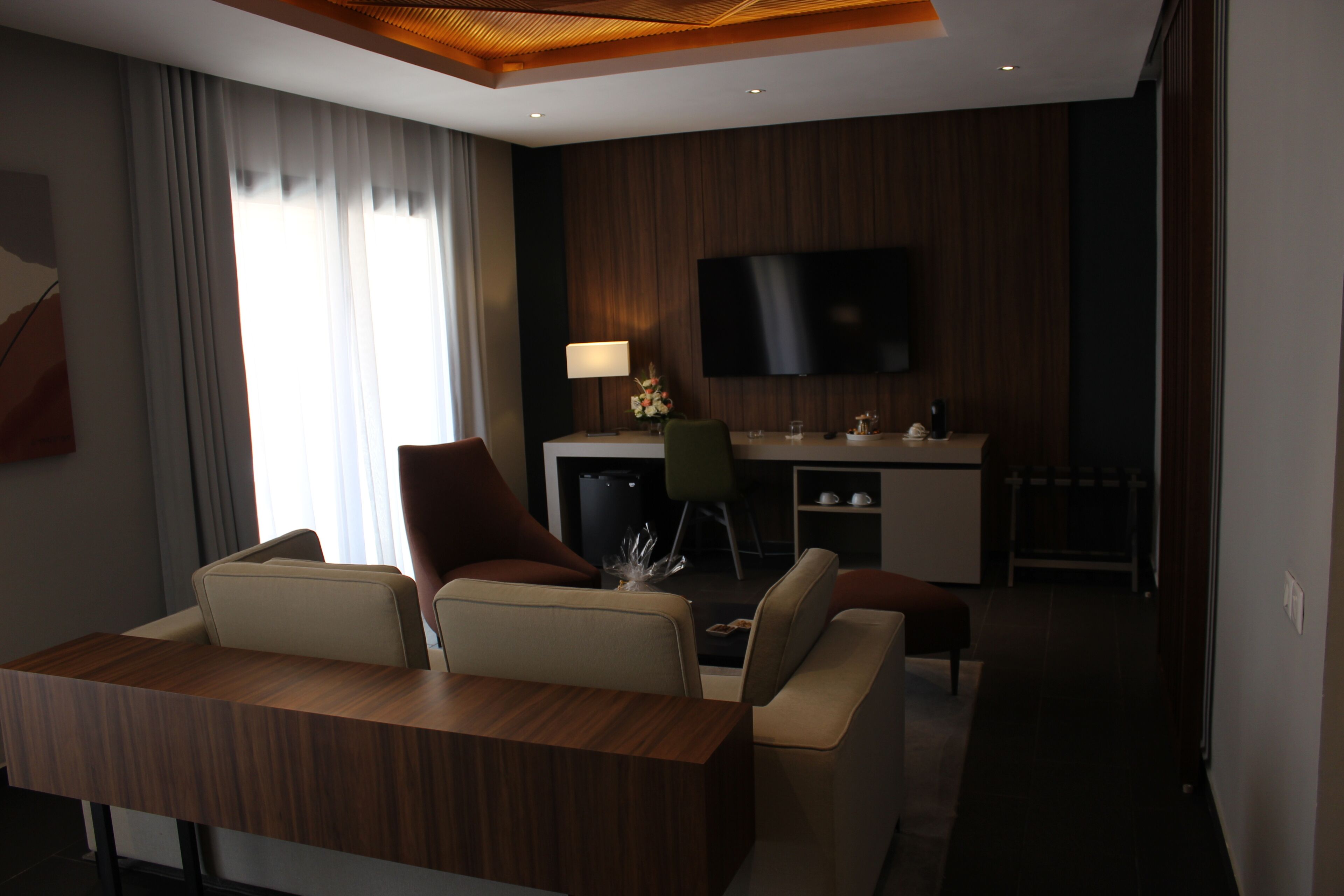 A modern hotel room with a wooden dining table, two sofas, a flat-screen TV mounted on the wall, and a small kitchenette area.