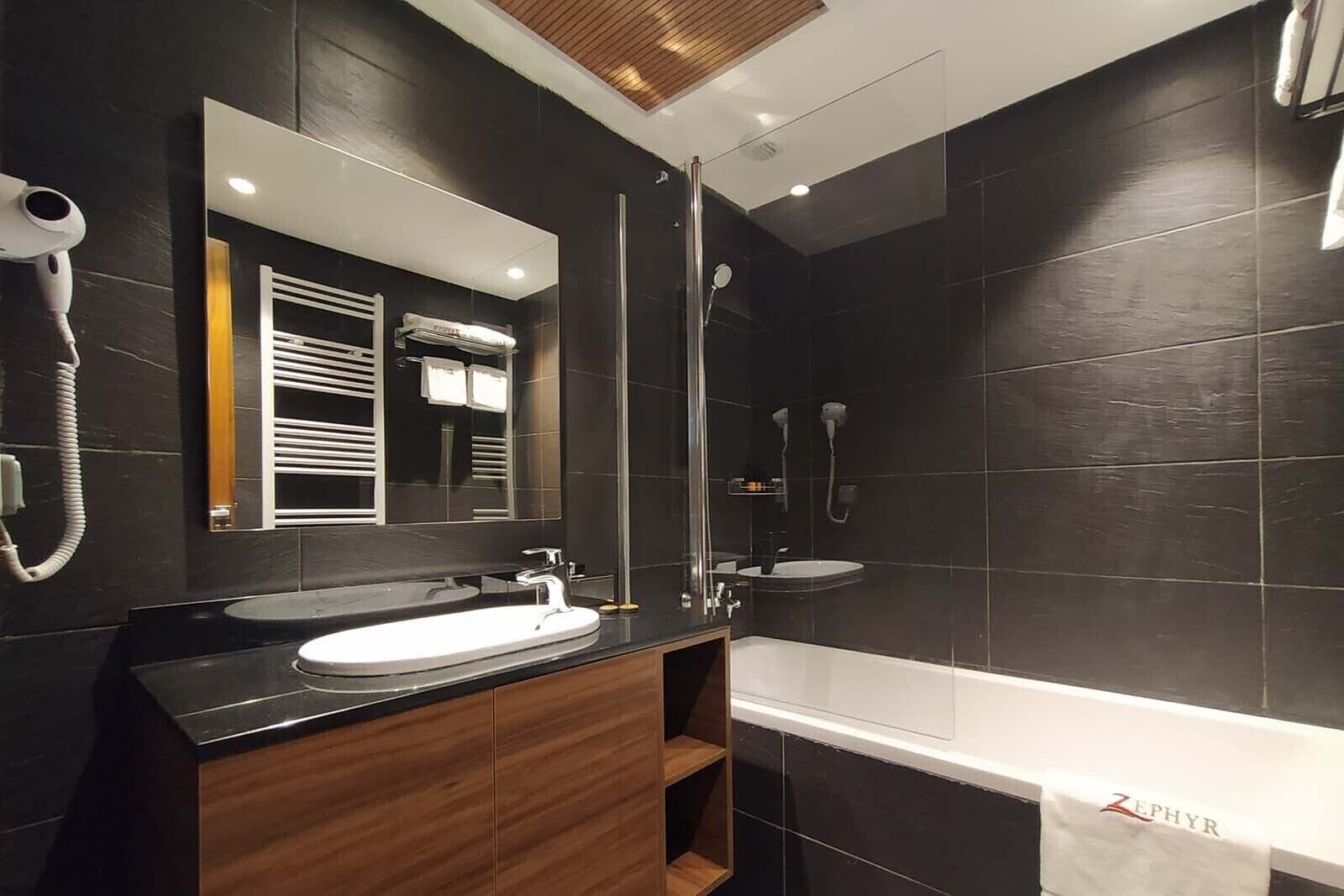Un baño moderno con baldosas oscuras, un mueble de tocador de madera, un espejo grande y una zona de ducha separada.