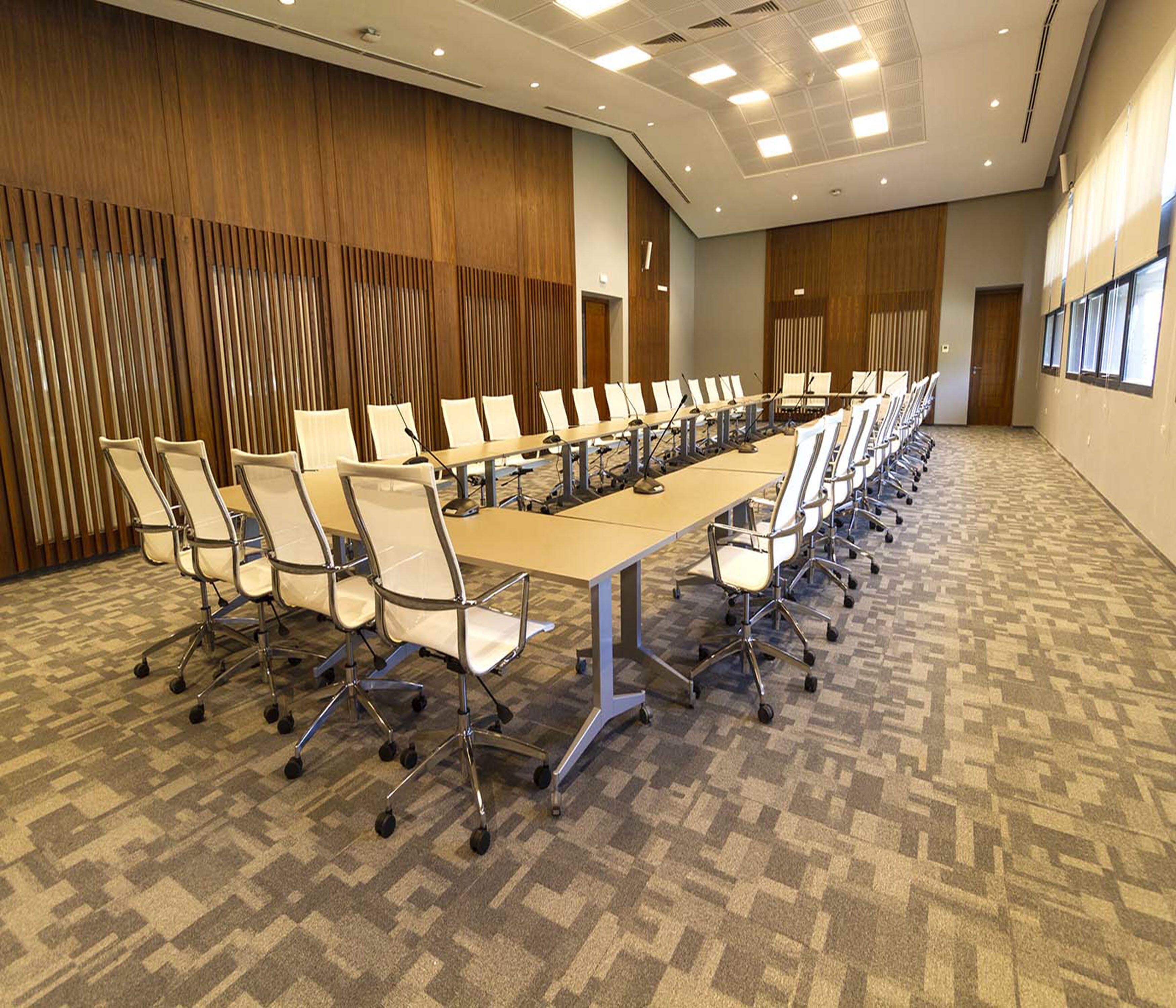 Un salón de conferencias con una larga mesa y sillas, suelo alfombrado, paneles de madera y luces en el techo.
