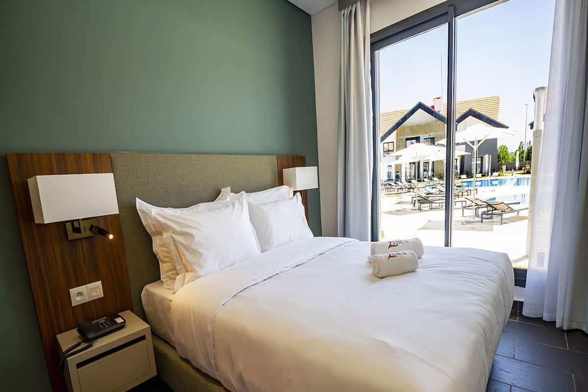 Habitación de hotel con una cama grande, una mesita de noche con teléfono y vistas a una piscina y mobiliario exterior.