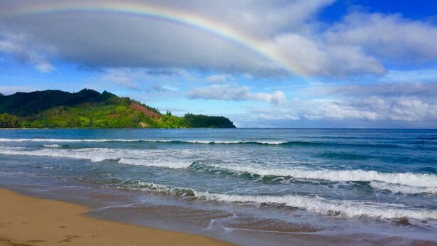 A beach with waves, a rainbow, and a green hill in the background. - Mamalahoa Mountain View Luxury Condo (Princeville)