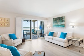 A living room with a white sofa, two armchairs, a coffee table, a balcony with outdoor furniture, and a painting on the wall.
