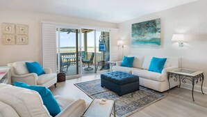 A living room with a sofa, ottoman, and chairs, a balcony with outdoor furniture, and a painting on the wall. - 1315 Pelican Watch Villas Beach & Free Amenities - Sale 249 USD (Seabrook Island)